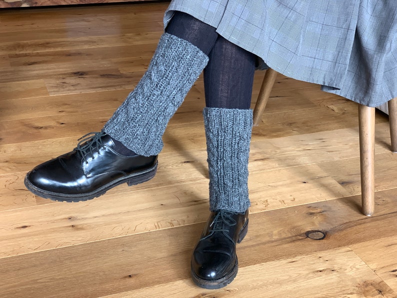 May include: Gray knitted leg warmers with a cable knit design are worn over black tights, paired with black leather shoes. The leg warmers add a touch of warmth and style to the outfit.