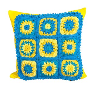 Könnte beinhalten: Ein gelbes Kissen mit einem blauen gehäkelten quadratischen Muster. Die Quadrate sind aus blauem und gelbem Garn gefertigt und haben ein blumenartiges Design in der Mitte.
