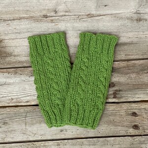 May include: A pair of green knitted arm warmers with a cable knit design. The arm warmers are made of a soft, textured yarn and are displayed on a weathered wooden surface. The cable knit pattern adds a decorative element to the warmers.
