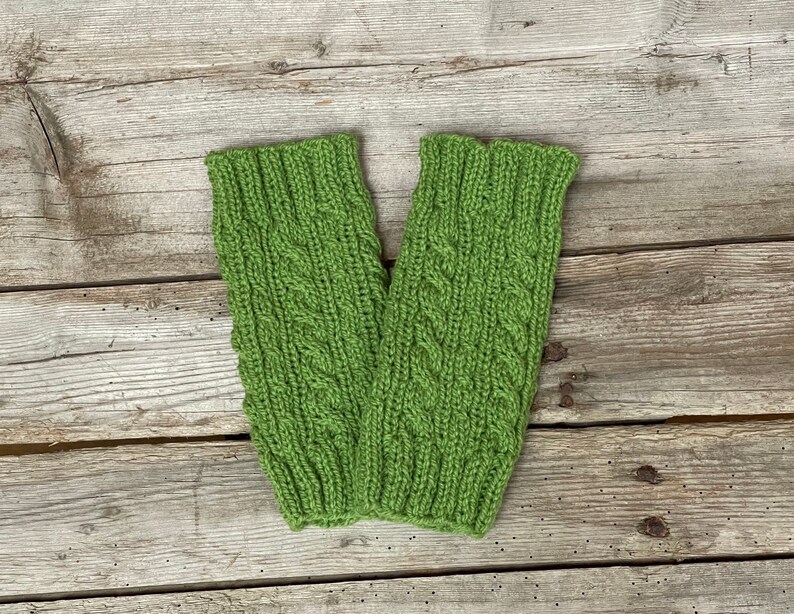 May include: A pair of green knitted arm warmers with a cable knit design. The arm warmers are made of a soft, textured yarn and are displayed on a weathered wooden surface. The cable knit pattern adds a decorative element to the warmers.
