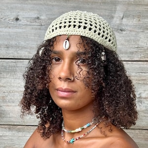 Può includere: Una persona indossa un cappello beige lavorato a uncinetto con una conchiglia e perline appese. Il cappello ha un design a uncinetto ed è indossato sulla testa. La persona ha i capelli castani ricci e indossa una collana con perline colorate.