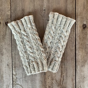Könnte beinhalten: Ein Paar beige gestrickte Armwärmer mit einem Zopfmuster. Die Armwärmer liegen flach auf einer Holzoberfläche.