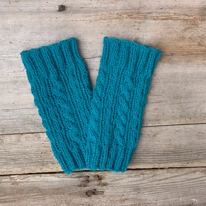 May include: A pair of teal-colored knitted leg warmers with a cable knit design. The leg warmers are made of a soft, textured yarn and are displayed on a weathered wooden surface. The ribbed cuffs and intricate pattern add a touch of handmade charm.