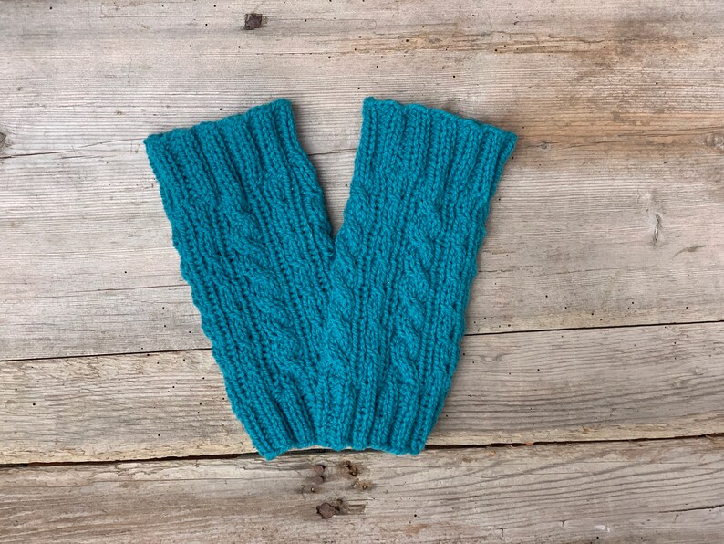 May include: A pair of teal-colored knitted leg warmers with a cable knit design. The leg warmers are made of a soft, textured yarn and are displayed on a weathered wooden surface. The ribbed cuffs and intricate pattern add a touch of handmade charm.