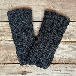 May include: A pair of dark gray knitted leg warmers with a cable knit design. The leg warmers are made of a soft, textured yarn and are displayed on a rustic wooden surface.