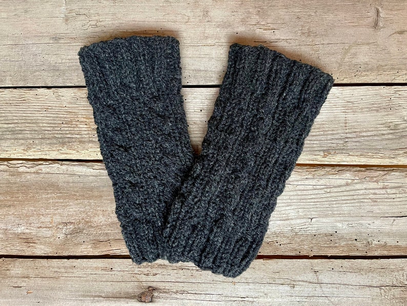 May include: A pair of dark gray knitted leg warmers with a cable knit design. The leg warmers are made of a soft, textured yarn and are displayed on a rustic wooden surface.