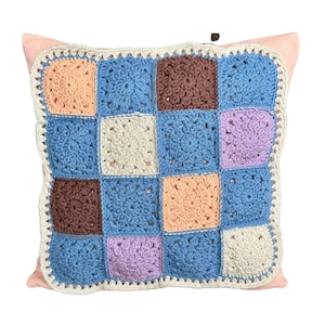 Könnte beinhalten: Ein quadratisches Häkelkissen mit Patchwork-Muster. Das Kissen besteht aus verschiedenen farbigen Quadraten, darunter blau, braun, rosa und beige. Das Kissen hat einen weißen Häkelrand.