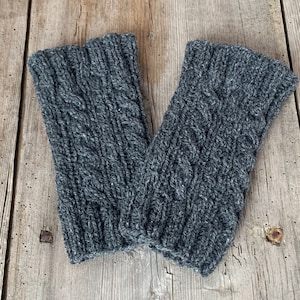 May include: A pair of gray knitted fingerless gloves with a cable knit design. The gloves are made of a soft, textured yarn and are designed to keep hands warm while leaving fingers free. The gloves are displayed on a wooden surface.