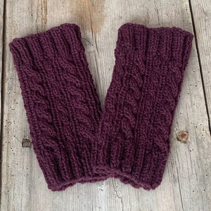 May include: A pair of knitted leg warmers in a deep burgundy color. The leg warmers feature a cable knit design and are made of a thick, textured yarn. They are displayed on a weathered wooden surface.