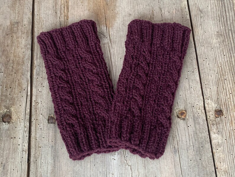 May include: A pair of knitted leg warmers in a deep burgundy color. The leg warmers feature a cable knit design and are made of a thick, textured yarn. They are displayed on a weathered wooden surface.