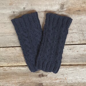 May include: A pair of navy blue knitted leg warmers. The leg warmers are made of a thick, textured yarn with a cable knit pattern. The cuffs are ribbed. The leg warmers are displayed on a wooden surface.