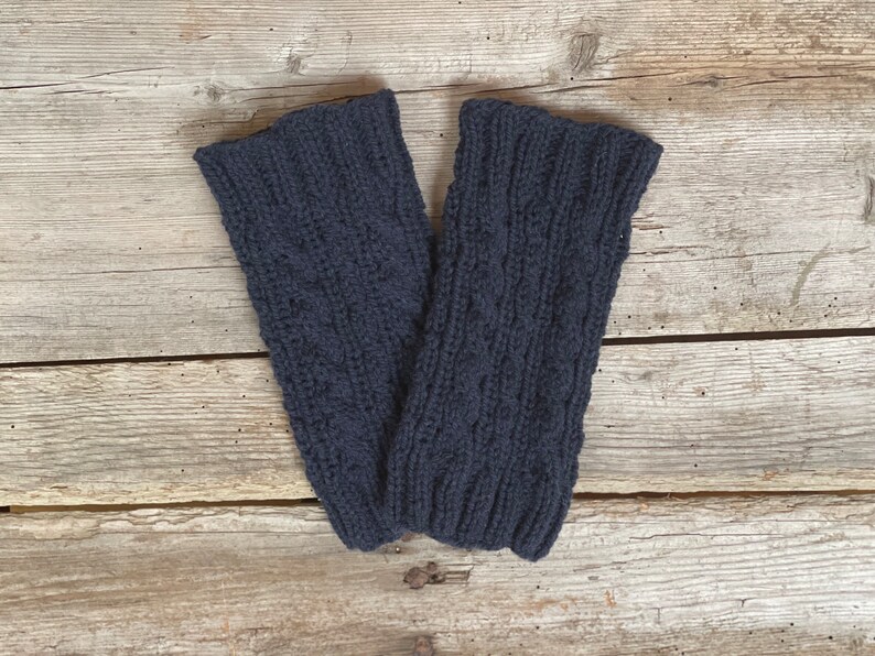 May include: A pair of navy blue knitted leg warmers. The leg warmers are made of a thick, textured yarn with a cable knit pattern. The cuffs are ribbed. The leg warmers are displayed on a wooden surface.