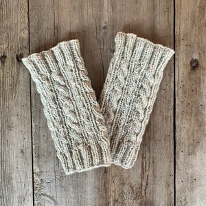 May include: A pair of light gray knitted arm warmers with a cable knit design. The arm warmers are made of a soft, textured yarn and are displayed on a weathered wooden surface. The cable knit pattern adds a touch of elegance.
