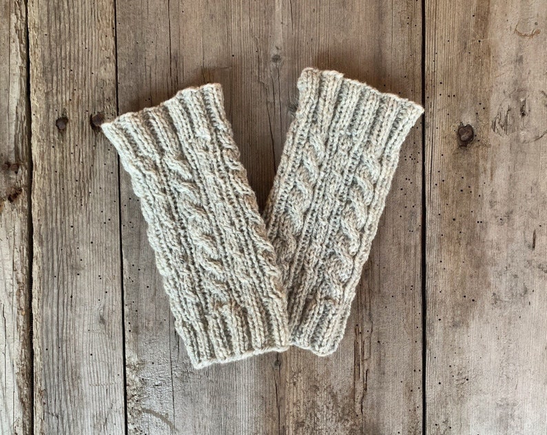 May include: A pair of light gray knitted arm warmers with a cable knit design. The arm warmers are made of a soft, textured yarn and are displayed on a weathered wooden surface. The cable knit pattern adds a touch of elegance.
