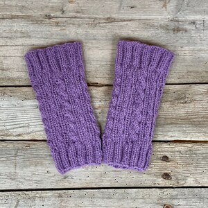 May include: A pair of lavender-colored knitted wrist warmers. The warmers feature a cable knit design in the center and ribbed cuffs. The background is a weathered wooden surface.