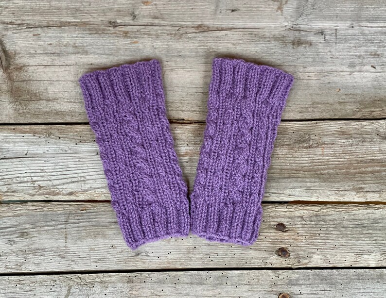 May include: A pair of lavender-colored knitted wrist warmers. The warmers feature a cable knit design in the center and ribbed cuffs. The background is a weathered wooden surface.