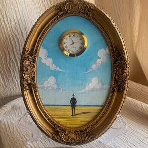 Könnte beinhalten: Ein ovales, goldfarben gerahmtes Kunstwerk mit einer surrealen Landschaft. Das Gemälde zeigt eine Figur, die an einem Strand unter blauem Himmel mit Wolken steht. Oben am Rahmen ist eine kleine Uhr angebracht. Der Rahmen hat Verzierungen.