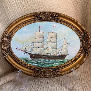 Puede incluir: Pintura ovalada enmarcada de un velero navegando sobre un mar verde bajo un cielo azul. El marco dorado adornado tiene detalles decorativos. El barco tiene velas blancas y banderas rojas. La pintura se exhibe sobre un soporte de plástico transparente.