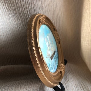 May include: A round, gold-colored decorative frame with ornate, raised detailing along the border. Inside the frame is a painting of a sailing ship on blue water under a light sky. The frame is supported by a small, black plastic display stand.