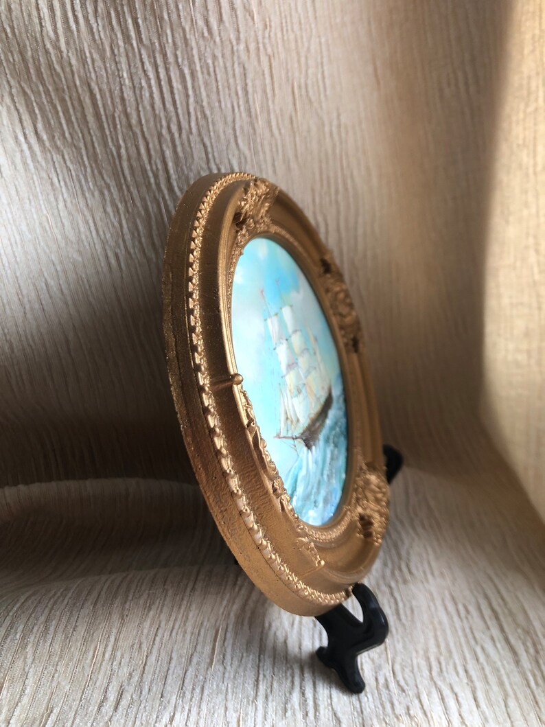 May include: A round, gold-colored decorative frame with ornate, raised detailing along the border. Inside the frame is a painting of a sailing ship on blue water under a light sky. The frame is supported by a small, black plastic display stand.