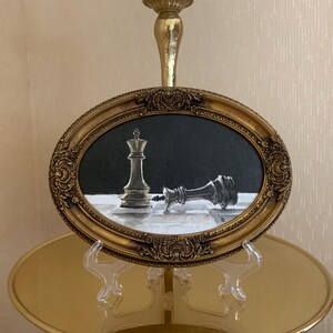 May include: An oval painting in an ornate gold frame depicting a chess scene with one upright king piece and one fallen king piece on a checkered board against a black background. The artwork features high contrast between the light and dark elements.