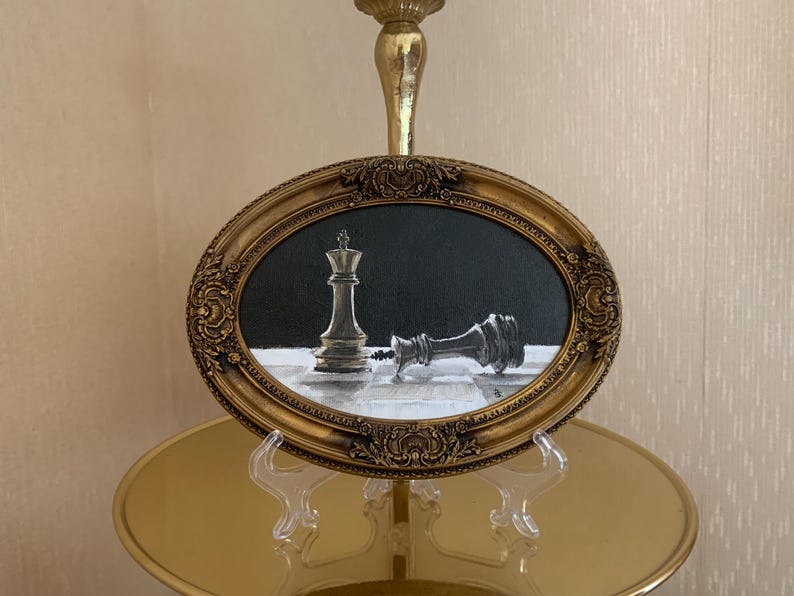 May include: An oval painting in an ornate gold frame depicting a chess scene with one upright king piece and one fallen king piece on a checkered board against a black background. The artwork features high contrast between the light and dark elements.