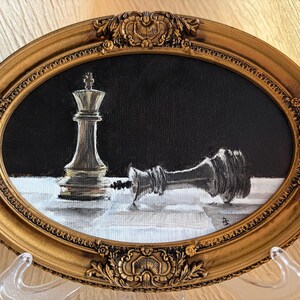 May include: An oval oil painting in a decorative gold frame featuring a standing white chess king and a fallen black chess king on a white surface against a black background. The painting captures the contrast between the two pieces in detail.