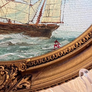 May include: An oil painting of a sailing ship on choppy green and white water, enclosed in an ornate, oval-shaped gold frame with intricate floral and scrollwork detailing. A red signature is visible on the lower right side of the canvas.