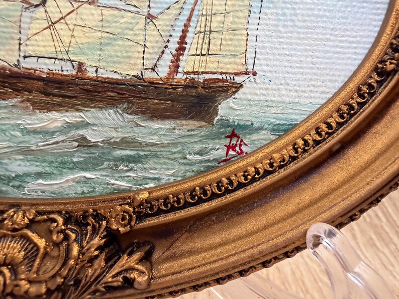 May include: An oil painting of a sailing ship on choppy green and white water, enclosed in an ornate, oval-shaped gold frame with intricate floral and scrollwork detailing. A red signature is visible on the lower right side of the canvas.