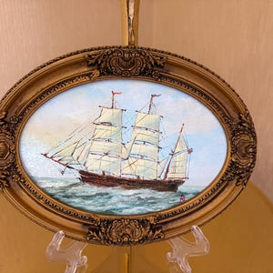 May include: An oval-shaped painting of a two-masted sailing ship on the ocean, enclosed in an ornate, metallic gold-colored frame with intricate relief detailing. The ship features white sails and a dark hull, set against a light sky.