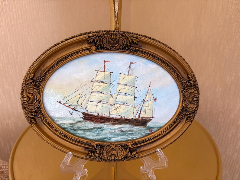 May include: An oval-shaped painting of a two-masted sailing ship on the ocean, enclosed in an ornate, metallic gold-colored frame with intricate relief detailing. The ship features white sails and a dark hull, set against a light sky.