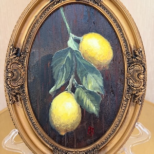 Pode incluir: Uma pintura oval em uma moldura dourada ornamentada apresentando dois lim&otilde;es amarelo-brilhantes pendurados em um caule verde com v&aacute;rias folhas verde-escuras contra um fundo marrom texturizado. Uma pequena assinatura vermelha &eacute; vis&iacute;vel no canto inferior direito.