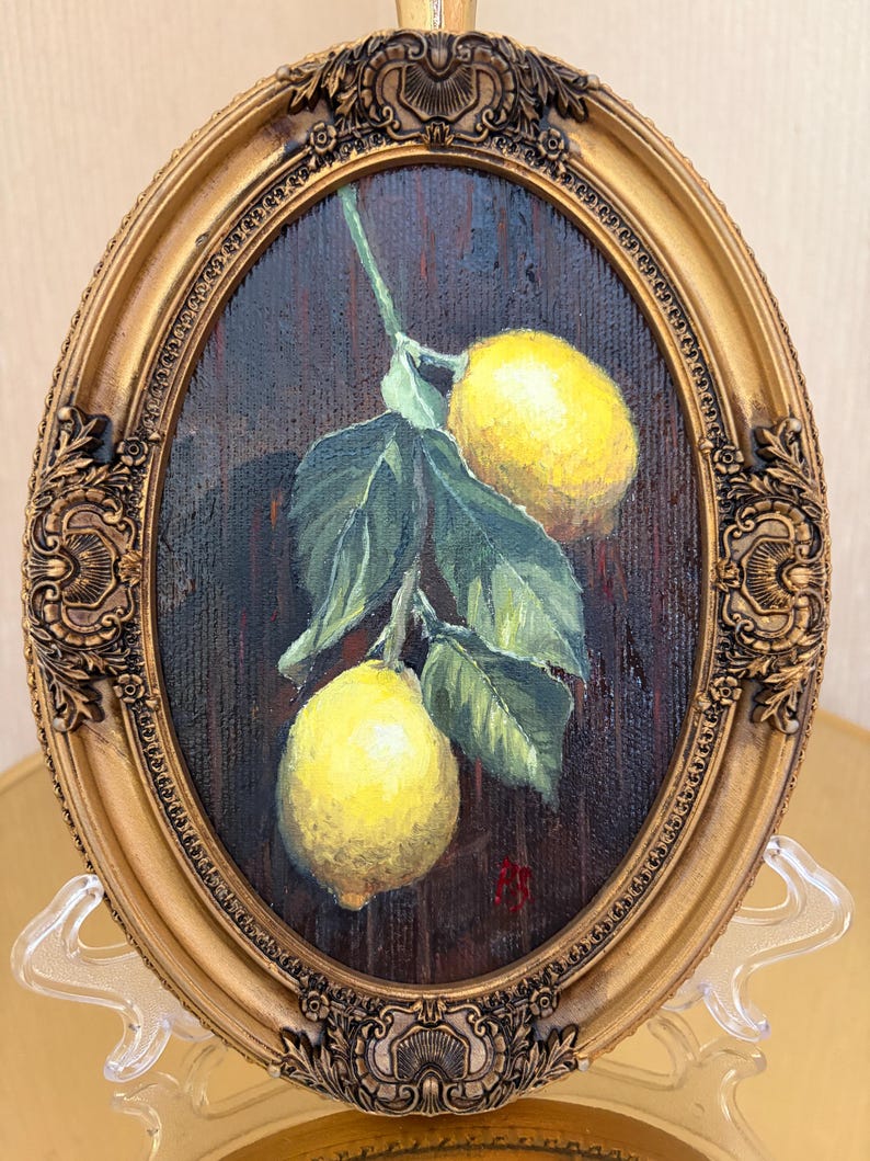 Pode incluir: Uma pintura oval em uma moldura dourada ornamentada apresentando dois lim&otilde;es amarelo-brilhantes pendurados em um caule verde com v&aacute;rias folhas verde-escuras contra um fundo marrom texturizado. Uma pequena assinatura vermelha &eacute; vis&iacute;vel no canto inferior direito.