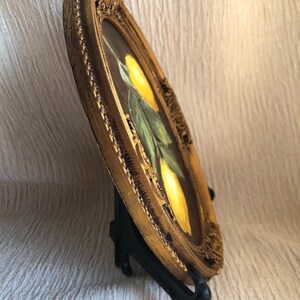 Pode incluir: Uma pintura de formato oval apresentando dois lim&otilde;es amarelos com folhas verdes sobre um fundo escuro, envolta em uma moldura dourada texturizada e ornamentada. A obra de arte &eacute; exibida em um suporte de pl&aacute;stico preto, vista de lado.