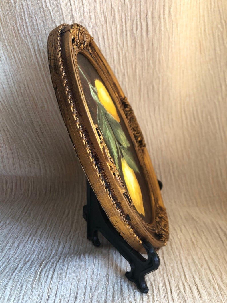 Pode incluir: Uma pintura de formato oval apresentando dois lim&otilde;es amarelos com folhas verdes sobre um fundo escuro, envolta em uma moldura dourada texturizada e ornamentada. A obra de arte &eacute; exibida em um suporte de pl&aacute;stico preto, vista de lado.