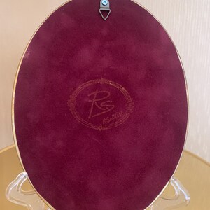 May include: An oval-shaped decorative plaque with a deep burgundy velvet backing and a gold-toned metal frame. The center features a circular embossed logo with the letters RS and the text R.SoillArt. A metal triangular hanging hook is attached.