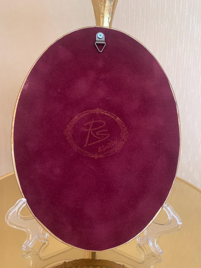 May include: An oval-shaped decorative plaque with a deep burgundy velvet backing and a gold-toned metal frame. The center features a circular embossed logo with the letters RS and the text R.SoillArt. A metal triangular hanging hook is attached.