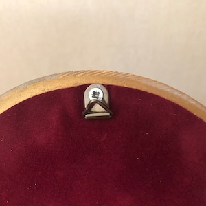 May include: The back of a circular object featuring a deep burgundy velvet-like fabric backing. A metallic triangular hanging hook is secured to the center top with a single silver-colored screw, framed by a smooth, light-toned wooden border.