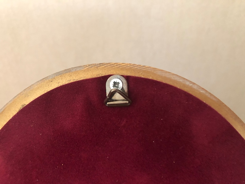 May include: The back of a circular object featuring a deep burgundy velvet-like fabric backing. A metallic triangular hanging hook is secured to the center top with a single silver-colored screw, framed by a smooth, light-toned wooden border.