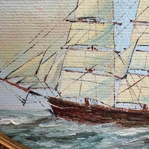 May include: An oil painting on canvas depicting a tall ship with brown wooden hull and cream-colored sails navigating through choppy, textured blue-green ocean waves. The artwork is enclosed within a decorative, ornate gold-toned oval frame.
