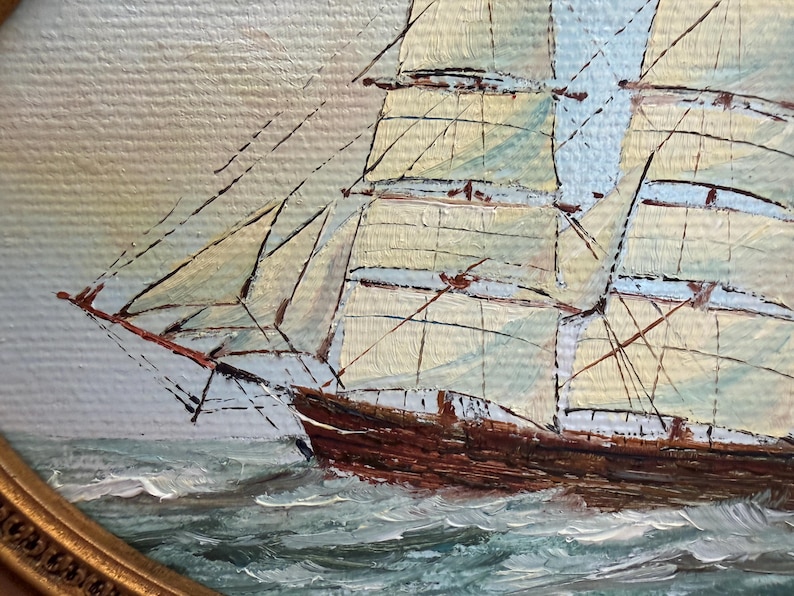 May include: An oil painting on canvas depicting a tall ship with brown wooden hull and cream-colored sails navigating through choppy, textured blue-green ocean waves. The artwork is enclosed within a decorative, ornate gold-toned oval frame.