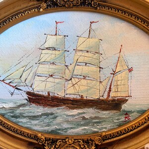 May include: An oval-framed oil painting depicting a brown sailing ship with white sails navigating choppy, dark green waters. The ship features multiple masts and rigging against a pale sky. The ornate gold frame has a textured, floral border.