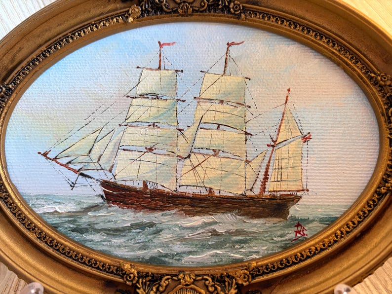 May include: An oval-framed oil painting depicting a brown sailing ship with white sails navigating choppy, dark green waters. The ship features multiple masts and rigging against a pale sky. The ornate gold frame has a textured, floral border.