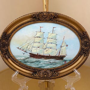 May include: An oval-shaped oil painting of a two-masted sailing ship on the ocean, featuring white sails and a dark hull. The artwork is encased in an ornate, metallic gold-toned frame with intricate relief detailing along the outer border.