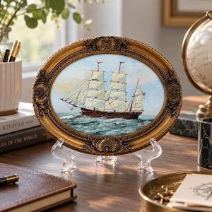 May include: An oval-shaped painting of a two-masted sailing ship on the ocean, enclosed in an ornate, antique-style gold frame with intricate relief detailing. The artwork depicts a brown hull with white sails against a light blue sky.