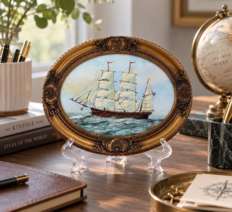 May include: An oval-shaped painting of a two-masted sailing ship on the ocean, enclosed in an ornate, antique-style gold frame with intricate relief detailing. The artwork depicts a brown hull with white sails against a light blue sky.