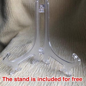 May include: A clear plastic display stand with a hinged design, featuring two curved support arms and a central vertical brace. The text "The stand is included for free (at your request)" is printed in red at the bottom of the image.
