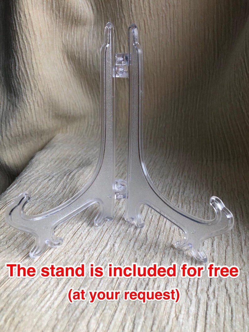 May include: A clear plastic display stand with a hinged design, featuring two curved support arms and a central vertical brace. The text "The stand is included for free (at your request)" is printed in red at the bottom of the image.