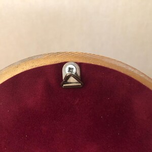 May include: A circular object with a smooth, light-colored wooden frame edge and a deep burgundy velvet backing. A small, silver-toned metal triangular hanging hook is securely fastened to the top center with a single visible metal screw.