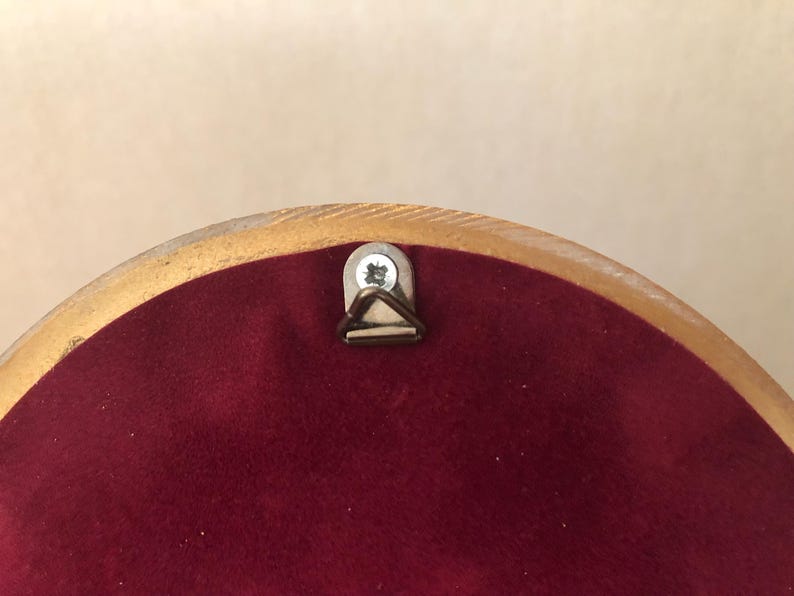 May include: A circular object with a smooth, light-colored wooden frame edge and a deep burgundy velvet backing. A small, silver-toned metal triangular hanging hook is securely fastened to the top center with a single visible metal screw.